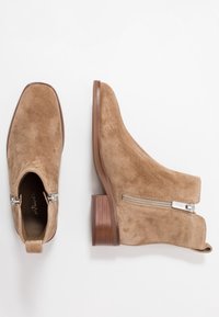 Beżowe zamszowe botki do kostki z niskim, drewnianym obcasem, wyposażone w boczny zamek i gładką fakturę. Projekt charakteryzuje się elegancką, nowoczesną sylwetką.