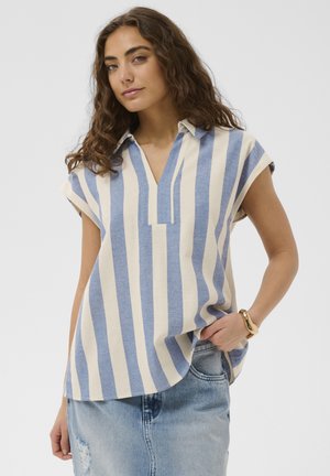 Jeune femme aux cheveux bouclés portant une chemise à manches courtes à rayures verticales bleues et blanches et une jupe en denim bleu clair, posant avec une main dans la poche.