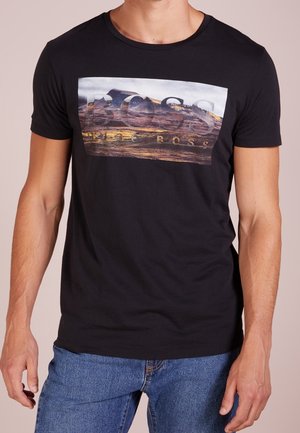 Musta puuvillainen t-paita, jossa graafinen painatus ja teksti "BOSS", lyhyet hihat ja pyöreä pääntie. Yhdistetty sinisiin farkkuihin.