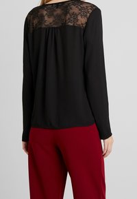 Blusa nera a maniche lunghe con un corpetto in pizzo trasparente, caratterizzata da una vestibilità ampia e dettagli plissettati sul retro, abbinata a pantaloni rossi.