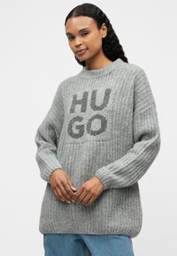 Gray knitted sweater with a ribbed texture, featuring the word "HUGO" in bold black letters on the front. Dropped shoulders and long sleeves.