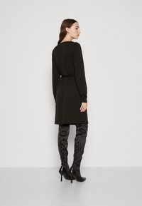 Svart knelang kjole med lange ermer, teksturert stoff og beltedetalj; sammen med høye, glitrende svarte støvletter og stiletthæler.