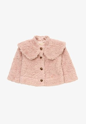 Ung pige iført en lyserød fluffy jakke, blomsterplisseret nederdel og gyldne sko, smilende og stående på en lys baggrund.