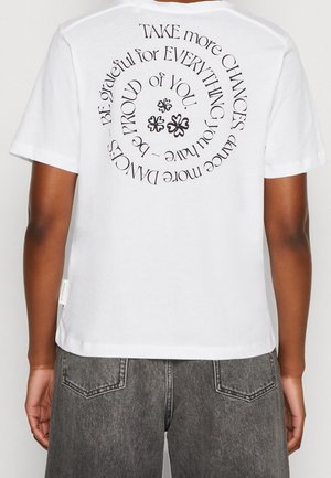 Person trägt weißes T-Shirt mit schwarzem, kreisförmigem Text auf dem Rücken, der lautet: "NIMM mehr CHANCEN, tanze mehr TÄNZE – SEI dankbar für ALLES, was du hast – Sei STOLZ auf DICH" mit kleinen Kleeblattsymbolen in der Mitte.