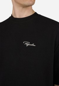 Juodas vaflinis marškinėlis su giliu apykaklę ir baltu siuvin�ėtu "Pegador" logotipu ant krūtinės. Tekstūruotas, laisvas kirpimas.