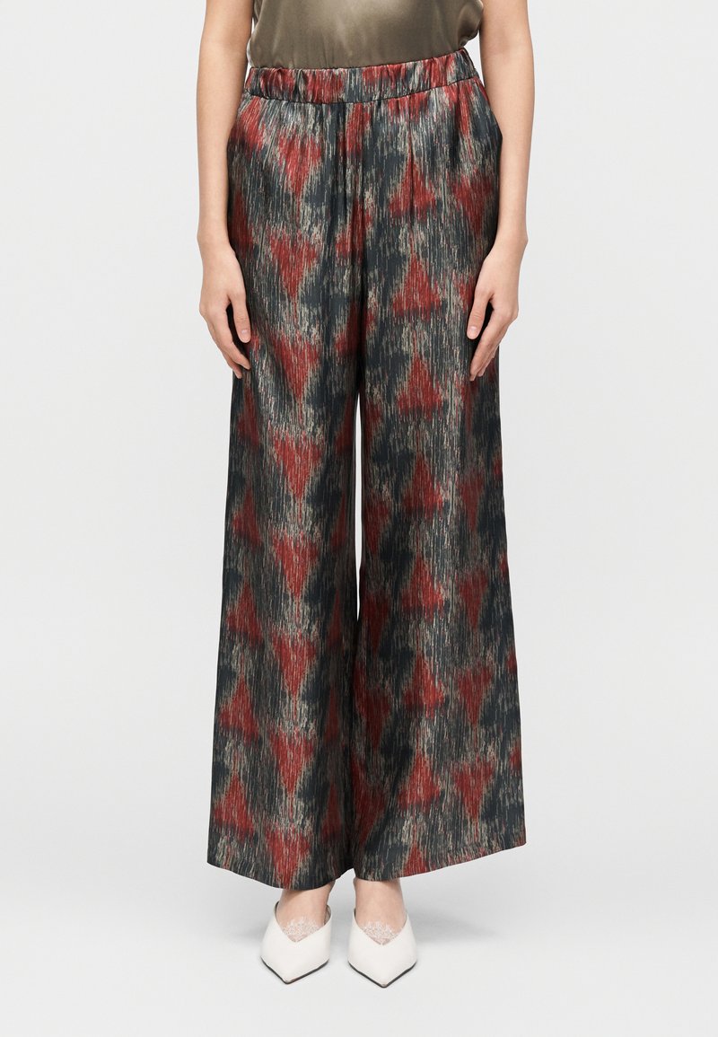 Calças de perna larga em tecido escuro com padrões geométricos vermelhos e cinzentos. Apresentam um cós elástico e textura suave. Combinadas com sapatos brancos.