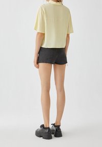Blouse à manches courtes, jaune, associée à un short en denim noir et des chaussures noires épaisses. Texture lisse avec un léger éclat sur le dessus.