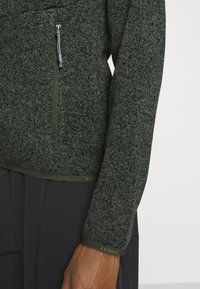 Dunkelgrüner Fleece-Pullover mit strukturiertem Finish, ausgestattet mit einer Reißverschlusstasche und gerippten Ärmeln. Der Saum hat eine kontrastierende Besatzdetail.