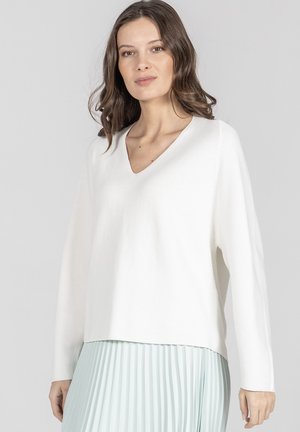 Kobieta z falującymi brązowymi włosami, ubrana w biały sweter z dekoltem w serek oraz jasnoniebieską plisowaną spódnicę, stojąca na jednolitym szarym tle.