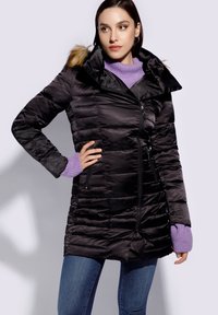 Abrigo de plumas negro con cierre de cremallera, que presenta una capucha forrada de piel, textura acolchada y bolsillos laterales. Combinado con un suéter púrpura y jeans.