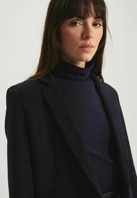 Blazer en tissu texturé bleu marine avec un col cranté soigneusement ajusté, porté sur un col roulé assorti. Des détails subtils rehaussent l'apparence sophistiquée.