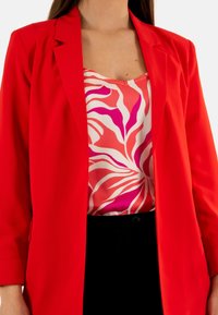 Blazer rojo con una textura suave, combinado con una blusa de estampado rosa y blanco que presenta diseños en espiral. Usado sobre tela negra.