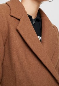 Abrigo de mezcla de lana marrón con solapa muesca, tejido texturizado y costuras visibles. Llevado sobre una sudadera con cremallera negra.