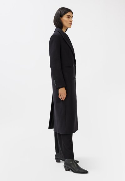 Cappotto nero in lana con silhouette lunga, colletto a falde, due bottoni e spacco laterale. Abbinato a pantaloni neri e stivaletti con punta.