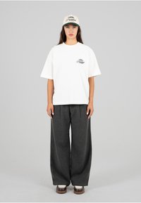 Hvit bomull T-skjorte med trykket logo, sammen med grå vidde bukser. Kappen er beige med en mørkegrønn skygge. Brunt fottøy fullfører looken.