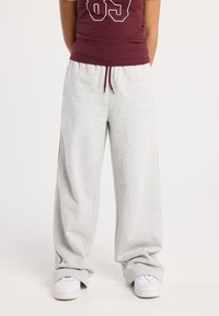 Personne portant un pantalon de survêtement large gris avec des rayures latérales, des baskets blanches et un débardeur bordeaux avec le numéro 85.