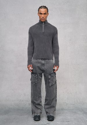 Suéter gris de canalé con cremallera en el cuello, combinado con pantalones cargo grises anchos que cuentan con grandes bolsillos y un ajuste relajado. Entorno: fondo de concreto minimalista.