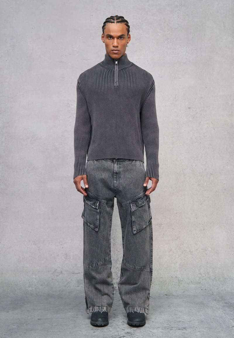 Suéter gris de canalé con cremallera en el cuello, combinado con pantalones cargo grises anchos que cuentan con grandes bolsillos y un ajuste relajado. Entorno: fondo de concreto minimalista.