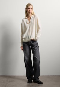 Un pull doux couleur crème avec un décolleté en V peu profond, associé à un jean en denim noir décontracté. Le mannequin porte des chaussures noires à enfiler.