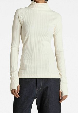 Frau trägt einen cremefarbenen, gerippten Langarm-Rollkragenpullover kombiniert mit dunkelblauen Jeans vor einem schlichten Hintergrund.