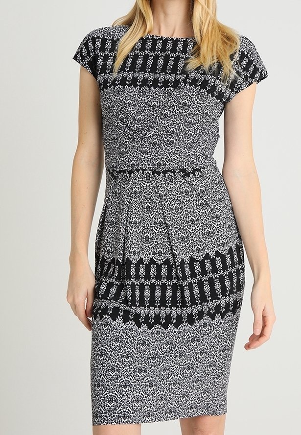 Femme portant une robe noire et blanche cintrée à motifs, longueur genou, avec des manches courtes et un col rond.