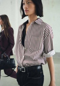 Camisa a rayas en burdeos y blanco, de manga corta, con un bolsillo. Combinada con jeans negros y un cinturón negro. El modelo se encuentra en un entorno minimalista.