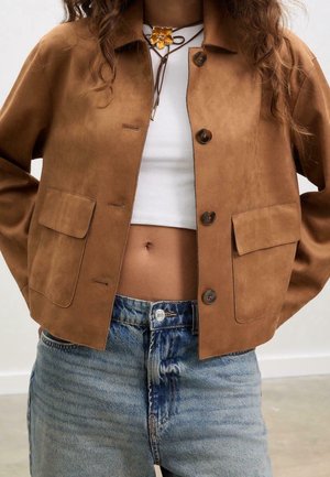 Frau trägt eine braune, geknöpfte Jacke mit großen Taschen, ein weißes Croptop, blaue Jeans und eine Halskette mit blumenförmigem Anhänger.