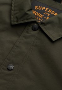 Zielona oliwkowa koszula robocza z usztywnionym kołnierzykiem, czarnymi guzikami i metką z pomarańczowym napisem. Gładki, trwały materiał z prostymi detalami w szwie.