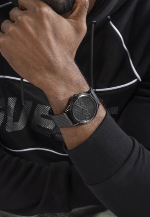Homme portant une montre-bracelet avec un bracelet en maille noire montrant le poignet et une partie de la main reposant près du menton, habillé d'un sweat à capuche noir avec des lignes blanches.
