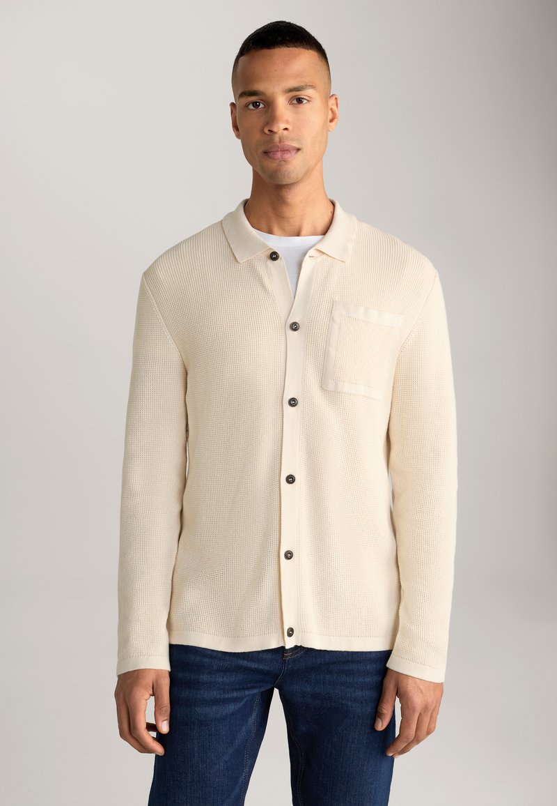 Cardigan beige lavorato a maglia con collare, chiusura con bottoni e una tasca sul petto. Presenta un tessuto testurizzato e maniche lunghe.