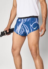 Blaue Sportshorts mit weißem abstraktem Muster, elastischem Bund und Nike-Logo. Person hält eine schwarze Wasserflasche.