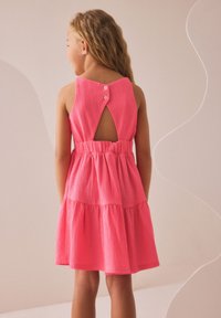 Różowa sukienka bez rękawów z falbanami, z wycięciem na plecach i dwoma guzikami. Wykonana z fakturowanej tkaniny.