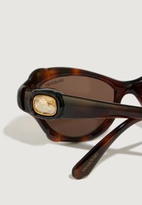 Óculos de sol em padrão tartaruga com lentes castanhas, apresentando uma armação preta com um detalhe de joia emrealçada a ouro na haste. Design leve em plástico.