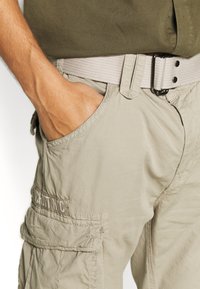 Pantalons cargo beiges en tissu texturé, avec des poches latérales et un écusson portant le texte "CLT INC." Accompagnés d'une ceinture texturée gris clair.