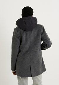 Abrigo de mezcla de lana gris con capucha de plumas negra desmontable, diseño ajustado, corte recto y aberturas laterales para facilitar el movimiento.