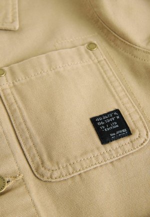Tissu kaki avec une finition texturée, présentant une poche avec du matériel en laiton et une étiquette noire indiquant les coordonnées et les détails de l'édition.