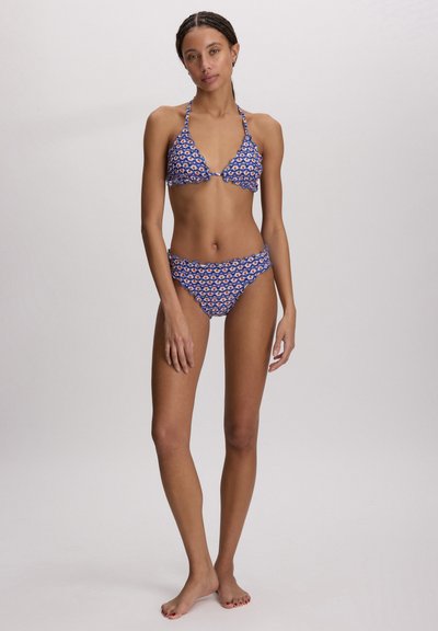 Kvinne stående barbeint iført en blåmønstret bikini med bølget kant mot en ensfarget lys bakgrunn.