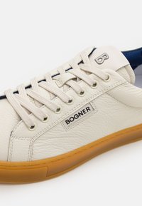 Hvit lær sneakers med teksturert finish, kremfargede lisser og en karmosin gummisåle. Har en logo og blå aksent nær hælen.