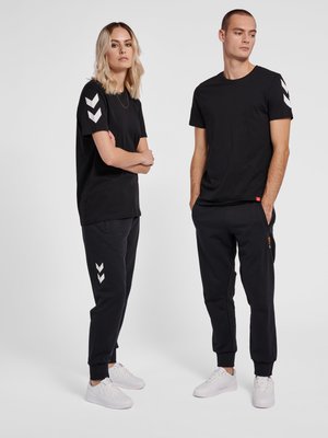 Due persone che indossano abiti casual neri con disegni a chevron bianchi sulle maniche e sui pantaloni, in piedi su uno sfondo chiaro e uniforme.