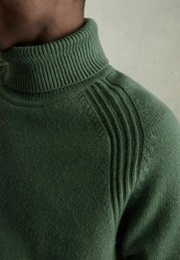 Pulover verde tricotat cu guler înalt cu nervuri, având accente cu nervuri pe umeri și o textură moale. Vedere de aproape a gulerului.