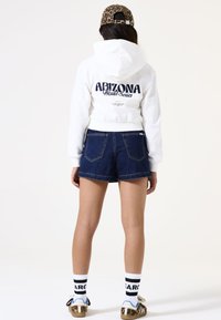 Sudadera recortada blanca con gráfico de "ARIZONA World Series"; pantalones cortos de mezclilla azules; gorra marrón con estampado; calcetas a rayas; y zapatos con patrón.