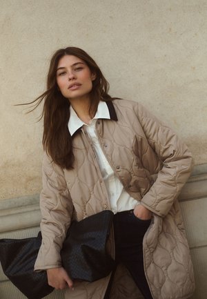Femme aux longs cheveux bruns portant un manteau matelassé beige par-dessus une chemise blanche, tenant une grande pochette noire texturée contre un mur en pierre.