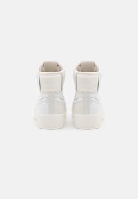 Valkoiset nahkakengät korkealla varrella ja pehmeillä beigeillä yksityiskohdilla, joissa on pehmustettu kaulus, ommeltu yksityiskohtia ja logo takana.