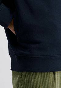Dunkelblaue Sweatshirt mit geripptem Saum, strukturiertem Stoff und Seitentaschen, kombiniert mit grünen Cordhosen.