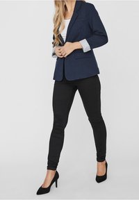 Blazer navy con maniche a righe arrotolate, abbinato a jeans skinny neri e tacchi alti neri. Tessuto morbido, design strutturato e vestibilità su misura.