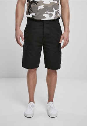 Mann trägt schwarze Cargo-Shorts, weiße Sneakers und ein graues Camouflage-T-Shirt und steht vor einem einfarbigen grauen Hintergrund.