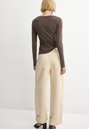 Donna con capelli biondi corti, indossa una maglia aderente a maniche lunghe marrone, pantaloni beige ampi a vita alta e scarpe con tacco nere, in piedi di spalle.