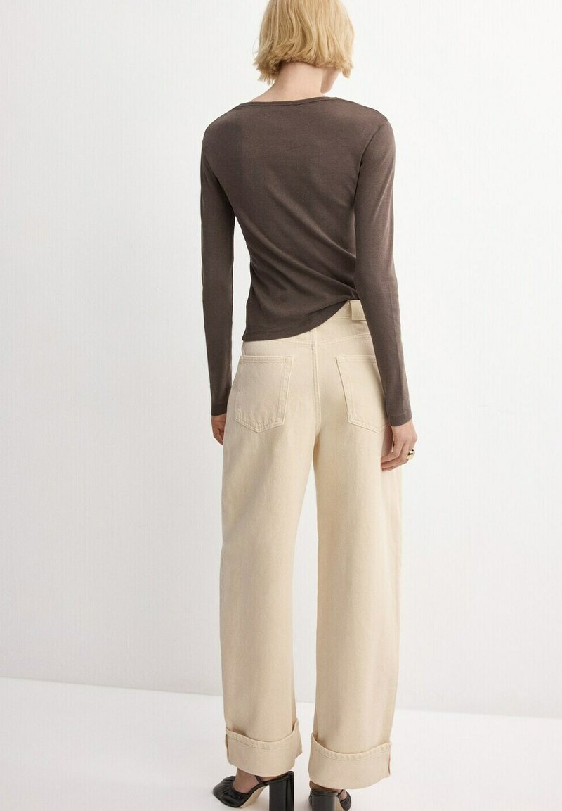 Femeie cu păr scurt blond, purtând un top maro cu mâneci lungi, pantaloni largi bej cu talie înaltă și pantofi negri cu toc, stând cu fața spre departe.