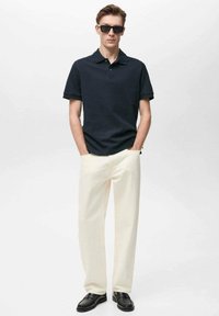 Námořnické polo tričko, krémové kalhoty a černé boty. Tričko má klasický límec, zatímco kalhoty jsou rovně střižené a s texturou.