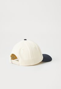 Baseball cap with a cream upper, black visor, and adjustable strap. Features ventilation holes and a round top button.
